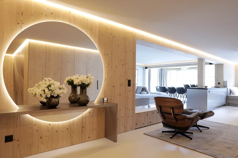 Interior of a modern room with wooden walls, large illuminated round mirror, flowers on a console, wooden chair, white flooring, and a dining table with chairs.