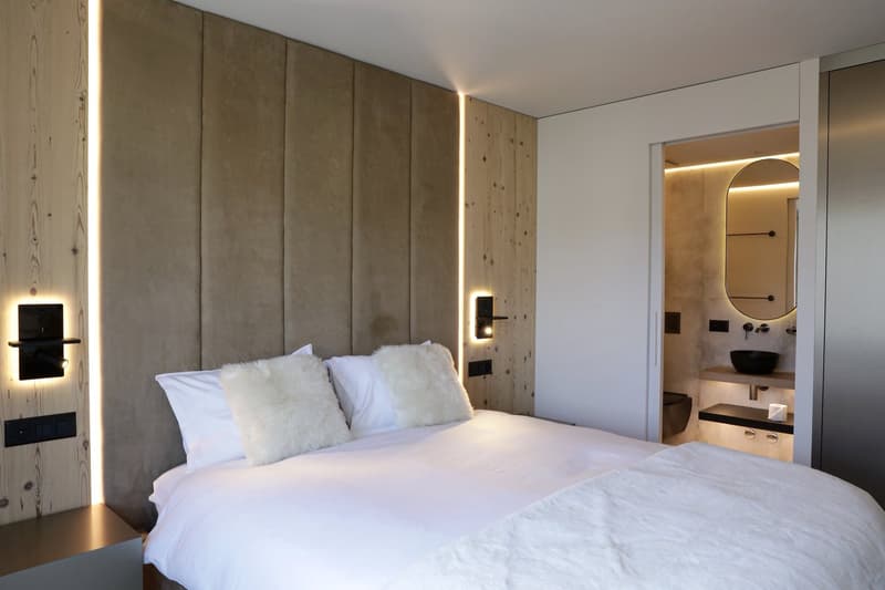 Bedroom with a bed, white linen, fluffy pillows, wooden wall design, wall mounted lights, open door to a washroom with sink, and toilet