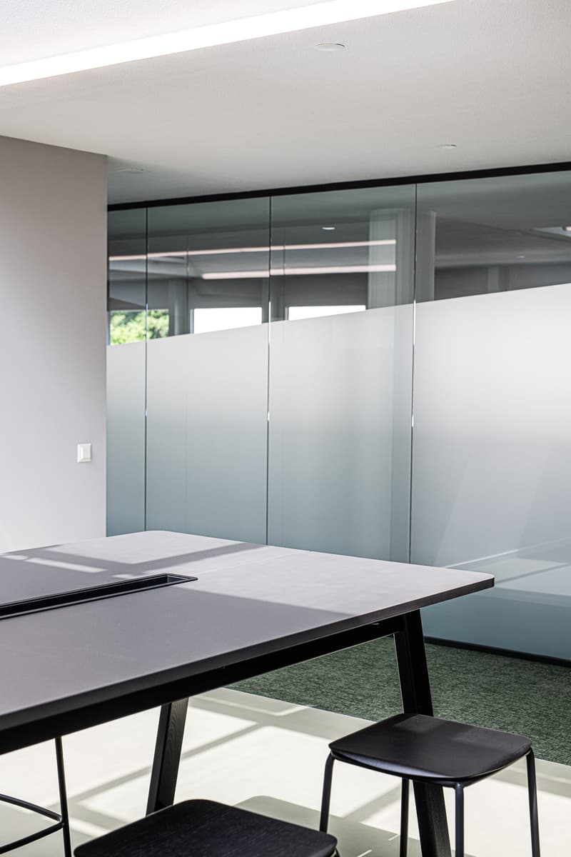 Glass office partition wall, black desk, black chairs, gray flooring, white ceiling, office light