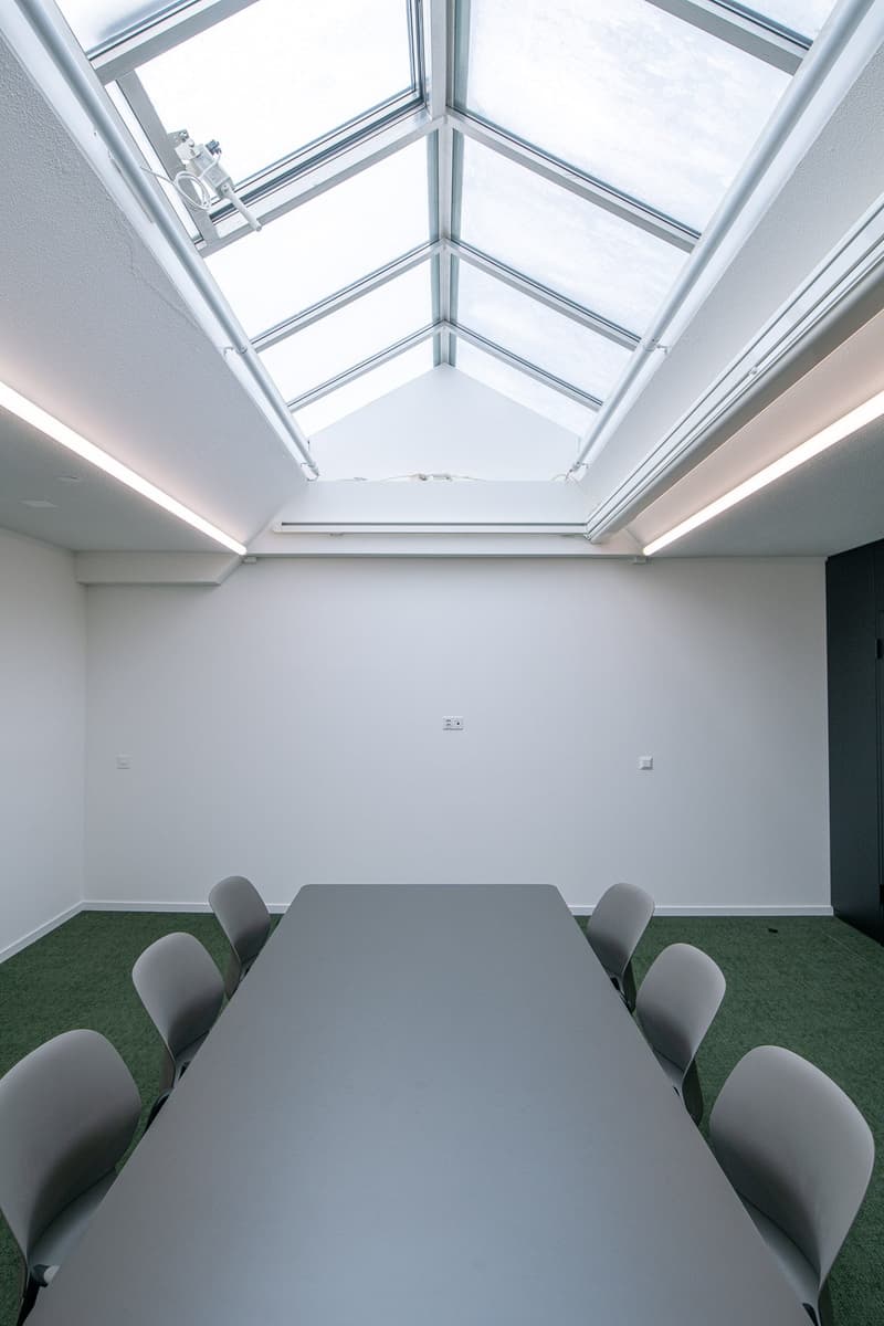 long rectangular conference table, 7 gray chairs, light gray ceiling with glass skylights, light gray walls