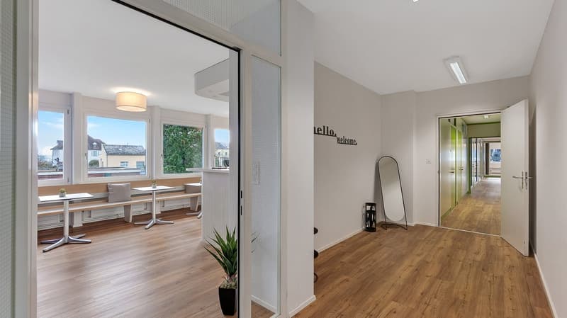 Open space with wooden floor, white walls, large windows, dining area with tables and benches, potted plants, hallway leading to another room