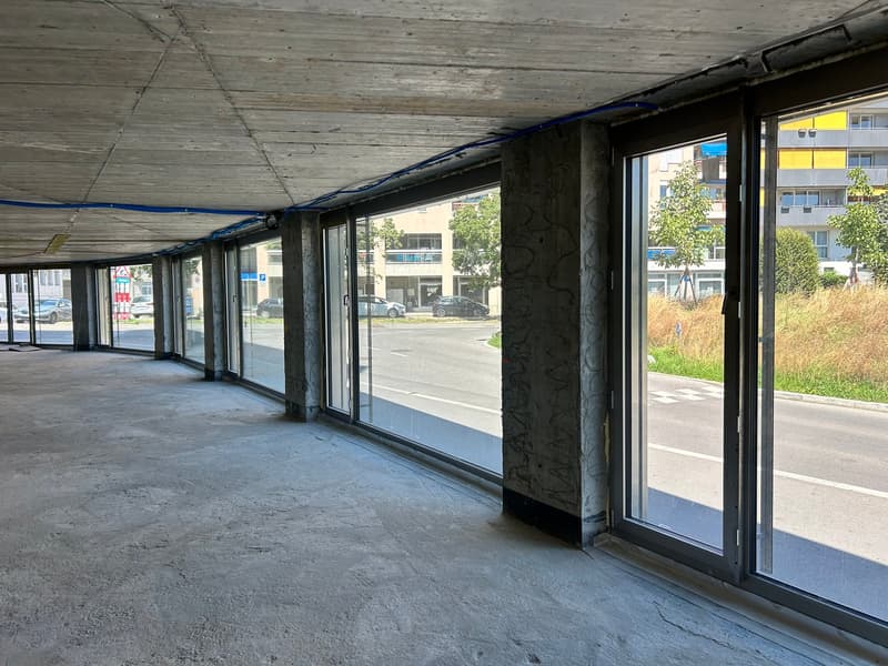 Large open commercial space with concrete floor, glass windows overlooking a parking lot, and a covered walkway or entrance area