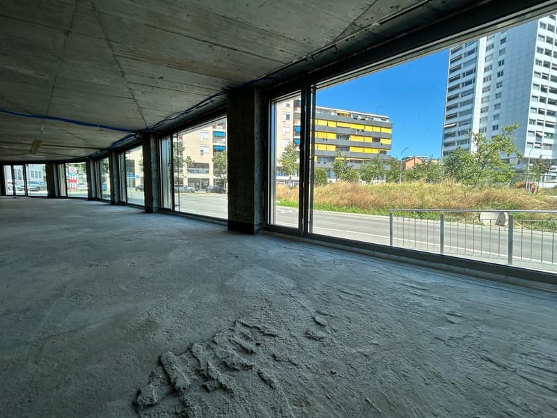 Large open space with concrete floor, glass windows overlooking the street and surrounding buildings, some of which have balconies or terraces visible.