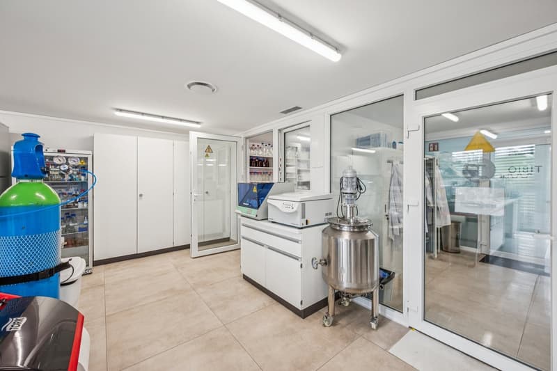 A laboratory with tile flooring, a large refrigerator, white cabinets, and industrial equipment such as a silver container and a cylindrical machine. The room has double-sided glass doors and is well-lit with overhead lights.