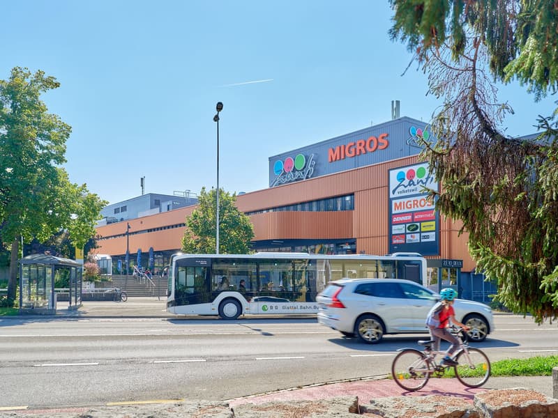 A large Migros supermarket building with a bus stop and people riding bicycles in front of it. The building has a distinctive orange and blue color scheme.