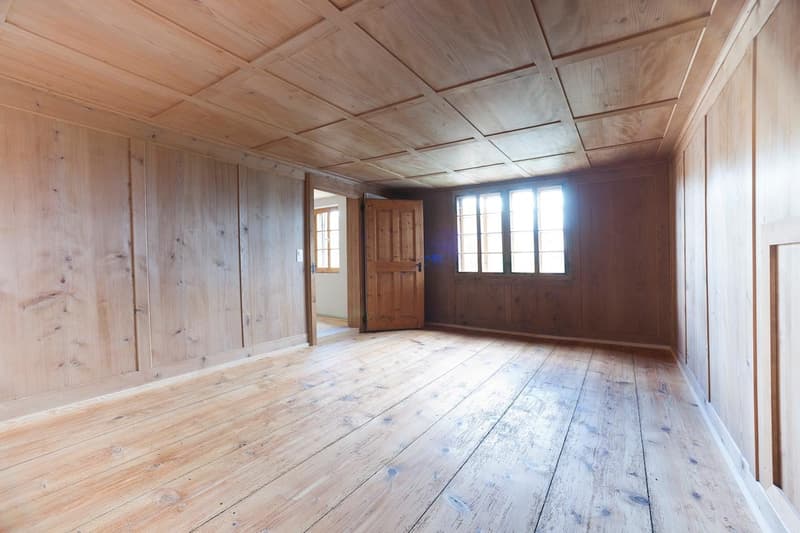 This image shows a large, open room with wooden walls and ceiling. The floor is made of distressed, wide wooden planks. There is a wooden door visible in the background, and several large windows that let in natural light. The overall aesthetic is rustic
