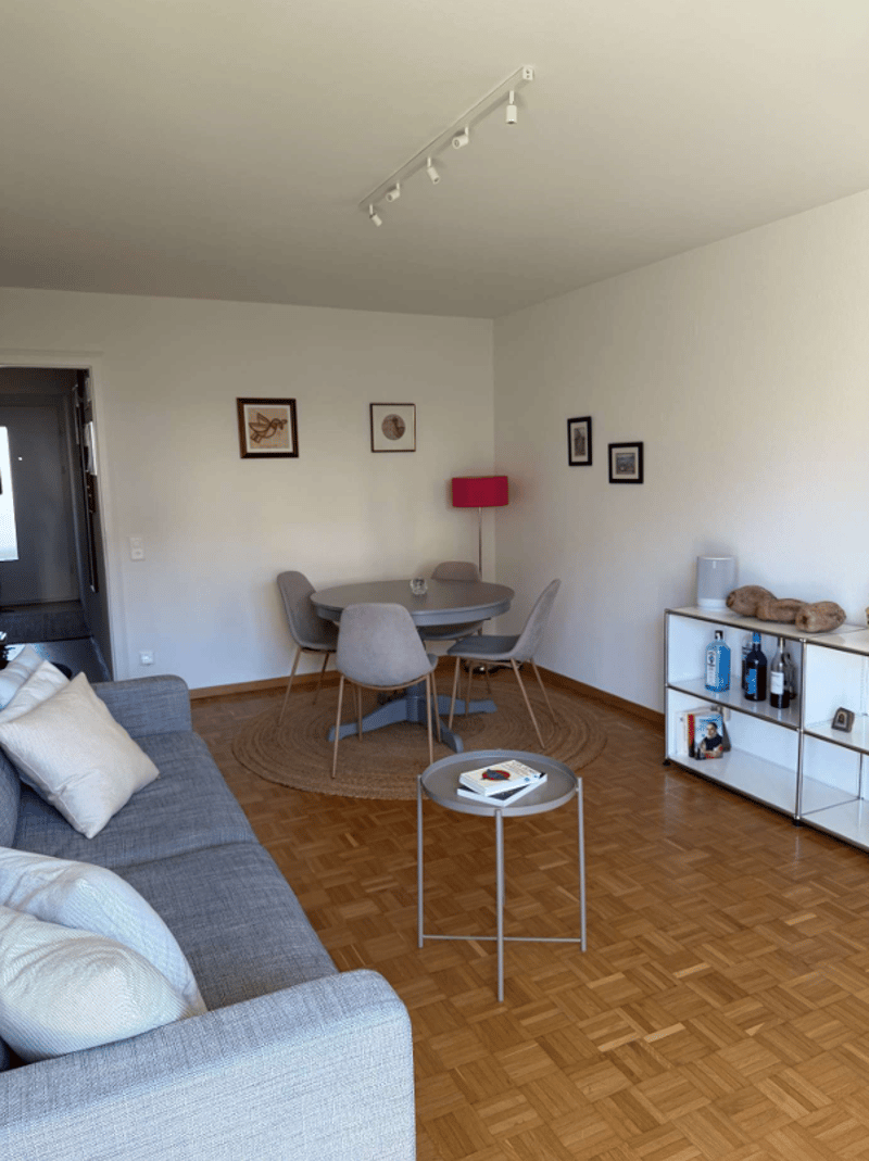 living room with wooden floor, gray couch, round table, round rug, round dining table, 4 chairs, lamp, shelves, framed pictures