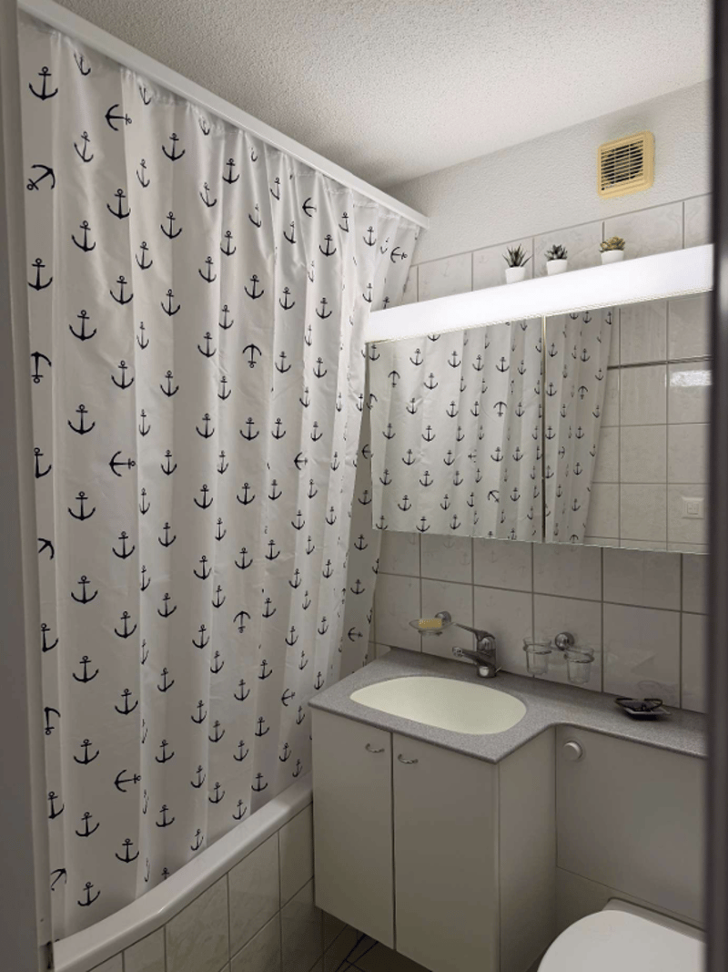Bathroom with white tiles, white shower curtain with anchor design, white sink, white toilet, white cabinet, mirror with light