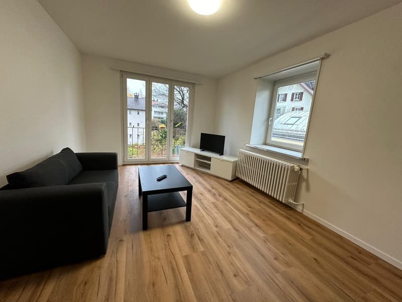 Spacious living room with hardwood flooring, large windows providing natural light, a black sofa, a coffee table, and a TV stand with a television.