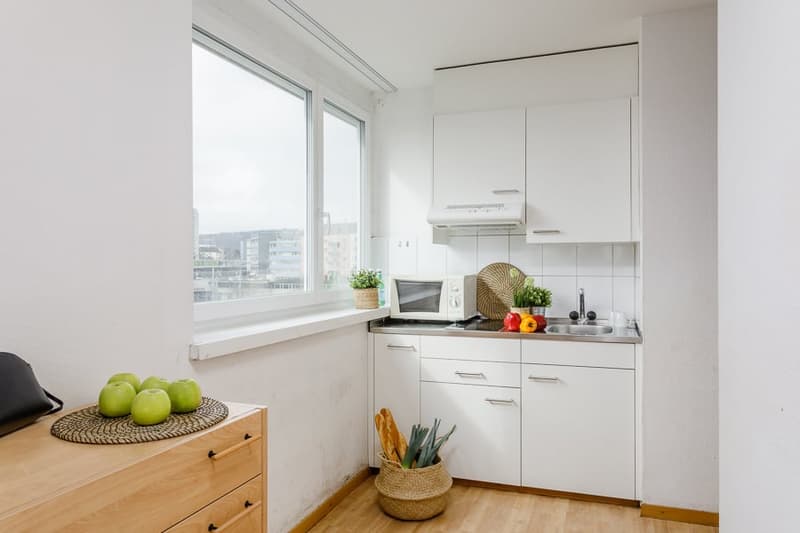 Compact kitchen with white cabinets, stainless steel appliances including a dishwasher, wooden countertop, and large window providing natural light