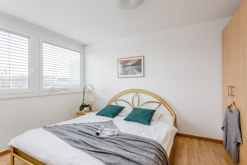 Spacious bedroom with large windows, white walls, hardwood floor, gold-framed bed, bedside table, and artwork on the wall