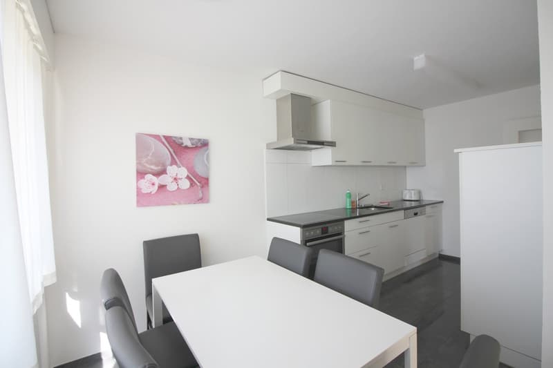 white tiled walls, kitchen with black countertop, white cabinets, modern appliances, grey chairs, dining table, artwork on wall, window with white curtains