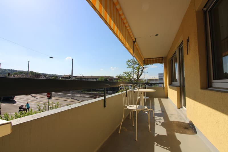 Outdoor balcony with a view of the city, two chairs and a small table, surrounded by a railing and overlooking a street with buildings and trees in the background.