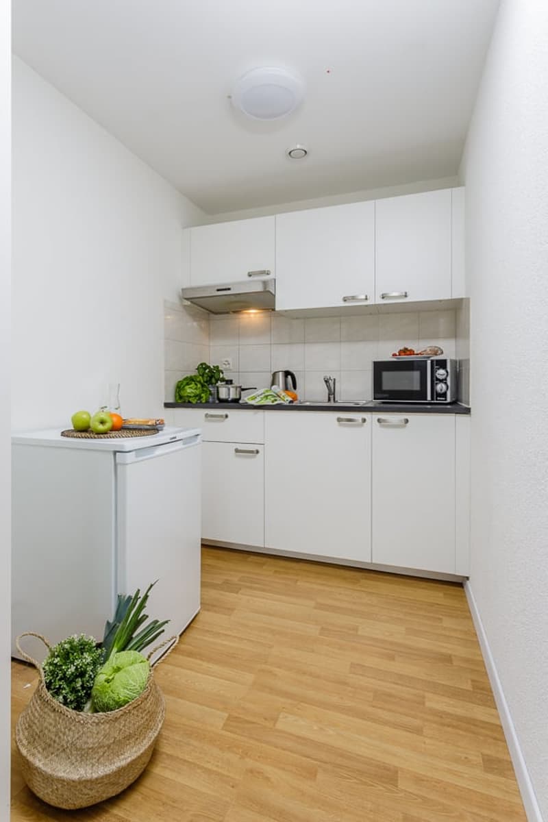 Compact kitchen with white cabinets, tiled backsplash, microwave, and refrigerator. Hardwood flooring and a basket with plants in the corner.