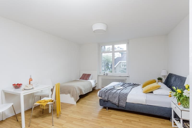 Spacious bedroom with hardwood flooring, white walls, large window, double bed, nightstand, and chair