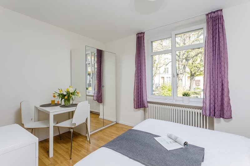 Bright and airy studio apartment with large windows, hardwood floors, white walls, a dining table, and purple curtains. The room has a couch and a mirror on the wall.
