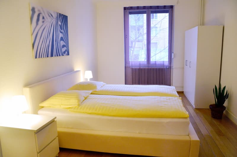 Bright and airy bedroom with white walls, large windows, a white bed with yellow bedding, a nightstand, and a wardrobe. The room also features a piece of artwork on the wall.