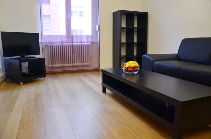 Spacious living room with hardwood floors, large windows, black TV stand, black bookshelf, blue sofa, and a coffee table with fruit on it.