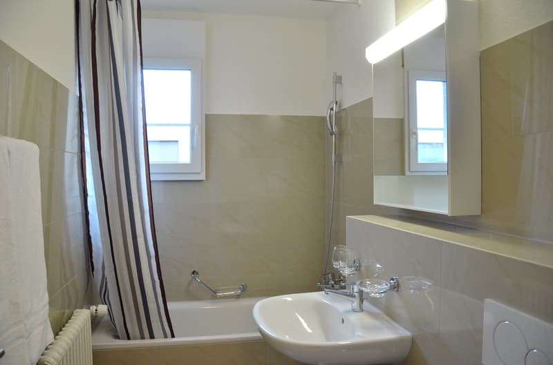 Tiled bathroom with a bathtub, sink, and shower curtain. The bathroom has a mirror and appears to be well-lit with natural light coming in through the windows.