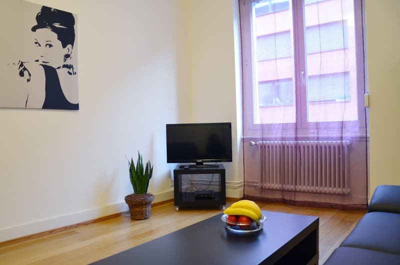 Spacious living room with hardwood floors, white walls, large windows, a TV stand with a TV, a potted plant, and a coffee table with fruit.