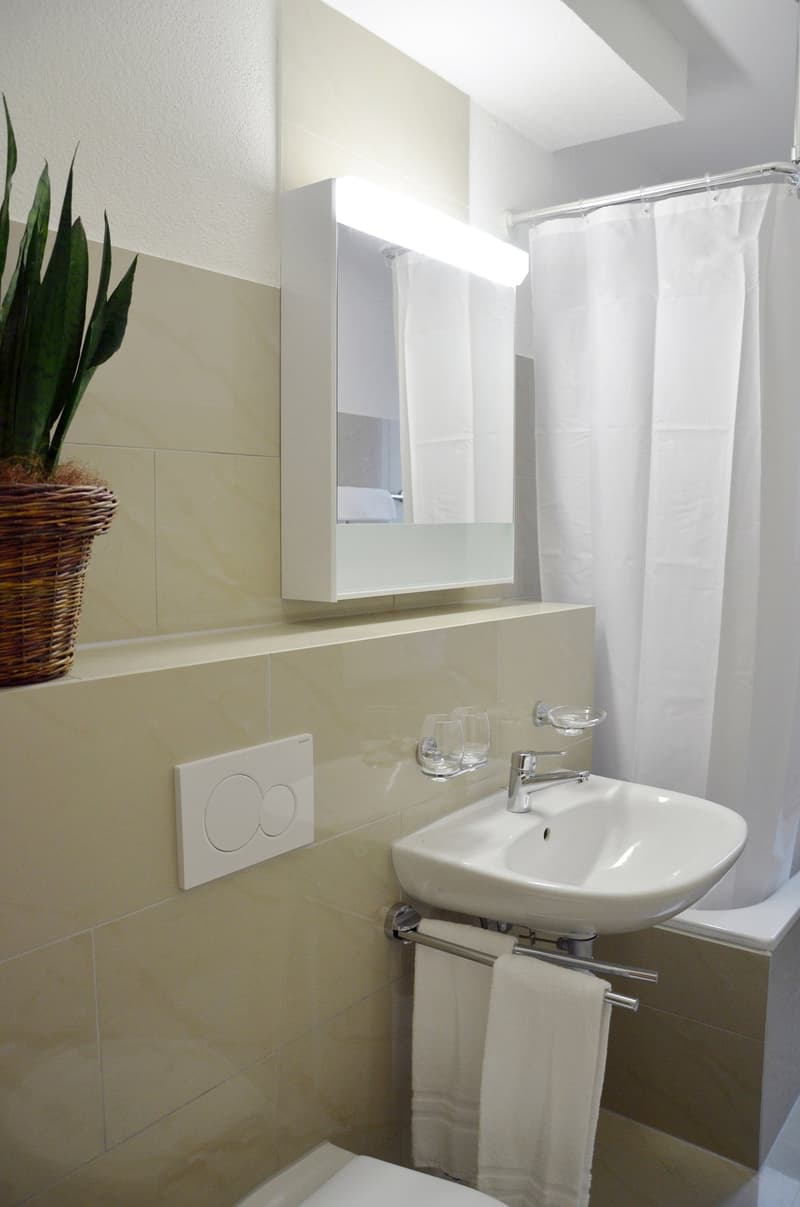 Bathroom with white tiles, a sink with a mirror cabinet above it, a shower curtain, and a plant in a wicker basket.