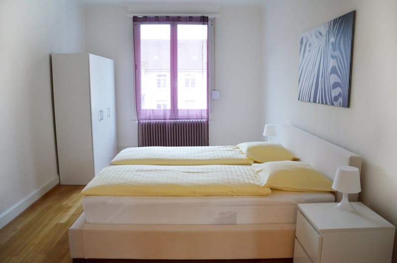 Spacious bedroom with white walls, hardwood floor, large window with purple curtains, white bed with yellow bedding, white wardrobe, and a modern artwork on the wall.