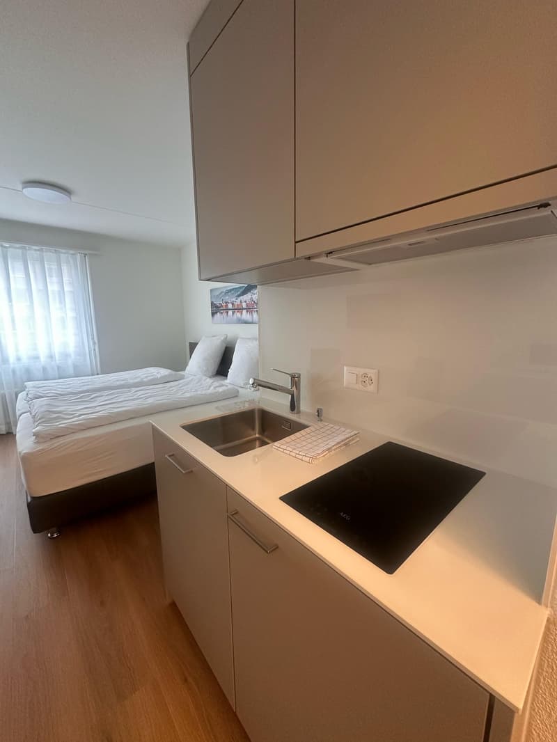 The image shows a studio apartment with a kitchenette and a sleeping area. The kitchenette features a sink, a cooktop, and built-in cabinets. The sleeping area has a bed and a small side table. The apartment has a clean, modern design with light-colored w
