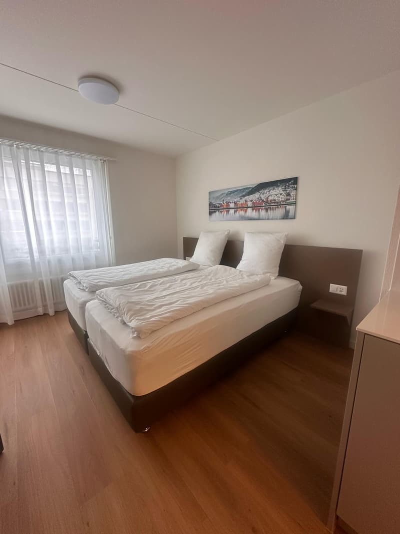 Spacious bedroom with hardwood flooring, white walls, large window, and a framed artwork on the wall. The room features a double bed with white bedding and a bedside table.