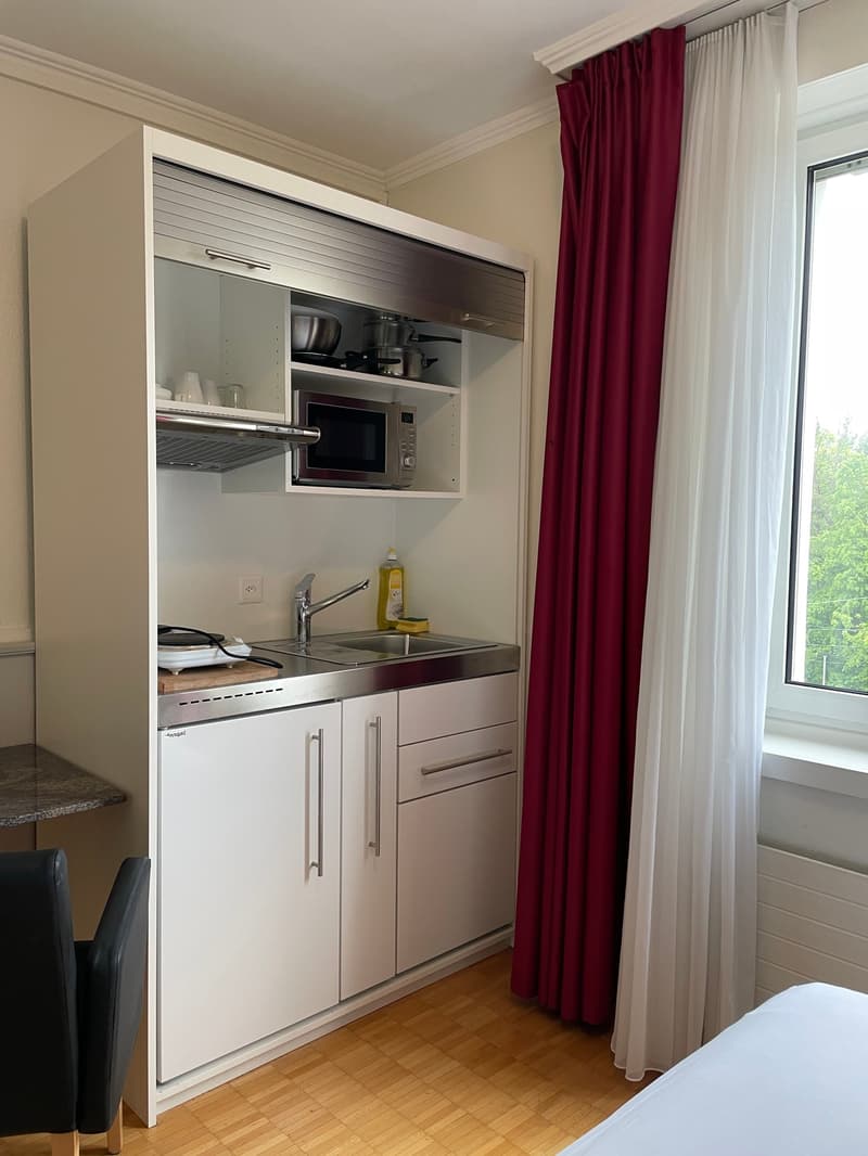 The image shows a compact kitchenette area within a room. It features a built-in cabinet with a microwave, storage space, and a small sink with a faucet. The kitchenette has a stainless steel finish and appears to be integrated into the room's design. The