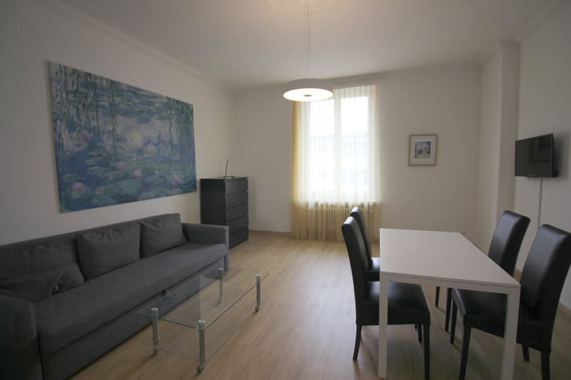The image shows a combined living room and dining room area. The living room features a gray sofa, a glass coffee table, and a large painting on the wall depicting a water lily pond. The dining room has a white table with four black chairs. There is a win