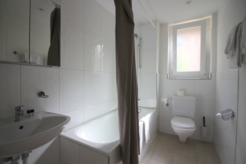 The image shows a modern bathroom with white tiles, a bathtub, a sink, and a toilet. The bathroom has a window that provides natural light. The overall design is clean and minimalist.