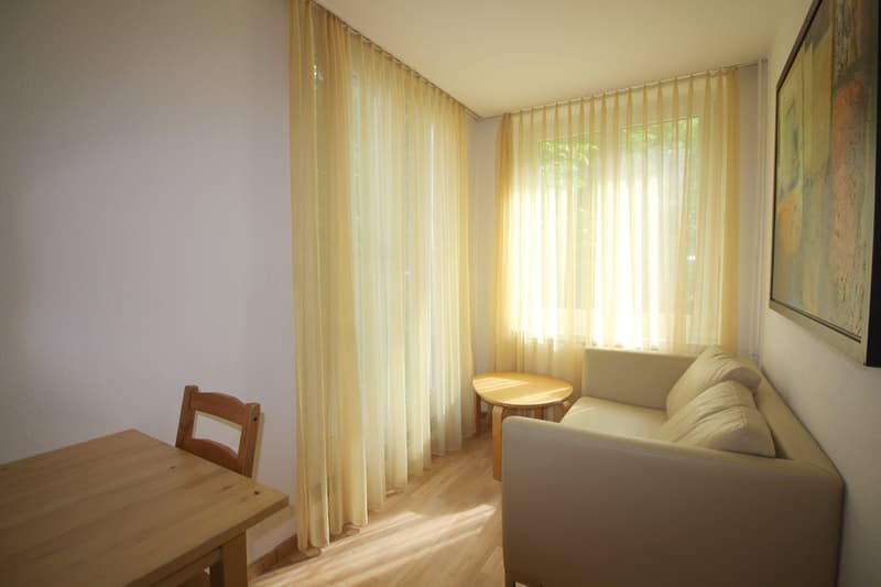 Spacious living room with large windows, beige curtains, wooden table and chair, and a comfortable sofa