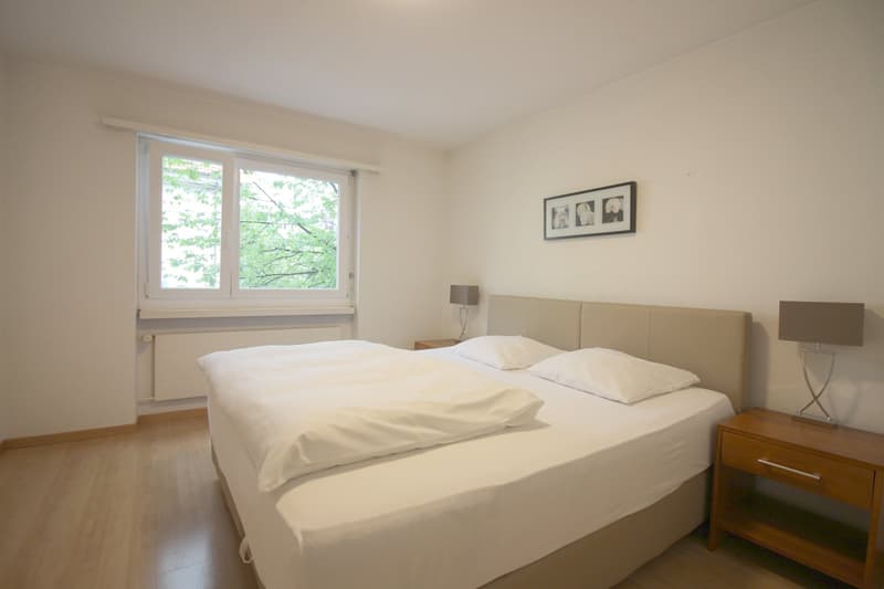 Spacious bedroom with large windows overlooking greenery, white walls, hardwood floors, a double bed with white bedding, a wooden nightstand, and a framed artwork on the wall.