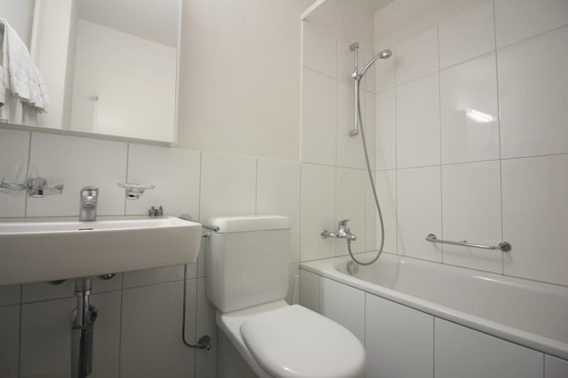 White tiled bathroom with a sink, toilet, bathtub, and shower head. The bathroom appears clean and modern.