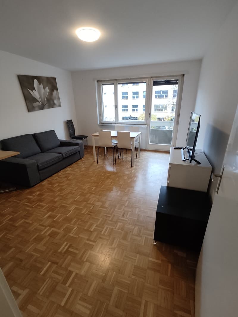 Spacious living room with hardwood flooring, large windows, a black sofa, a dining table with chairs, and a TV stand.