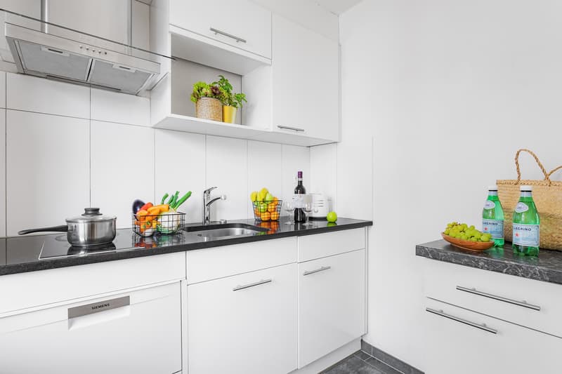 Modern, white kitchen with black countertops, stainless steel appliances including a dishwasher, and various kitchen accessories and produce on the countertop.