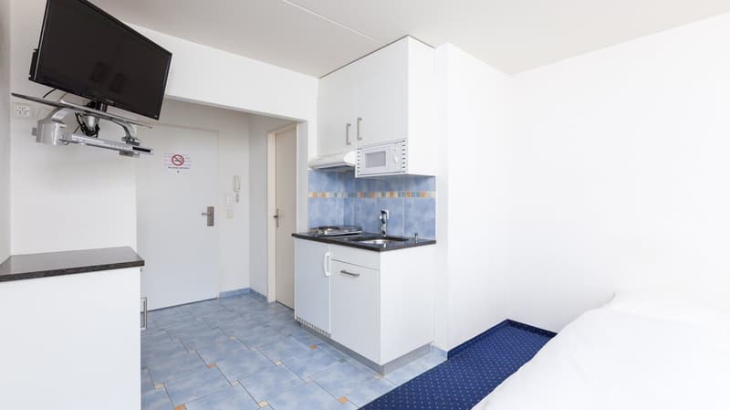 Kitchenette with sink, microwave, cabinets, and tiled floor, small bed, flat-screen TV on wall, and entrance door