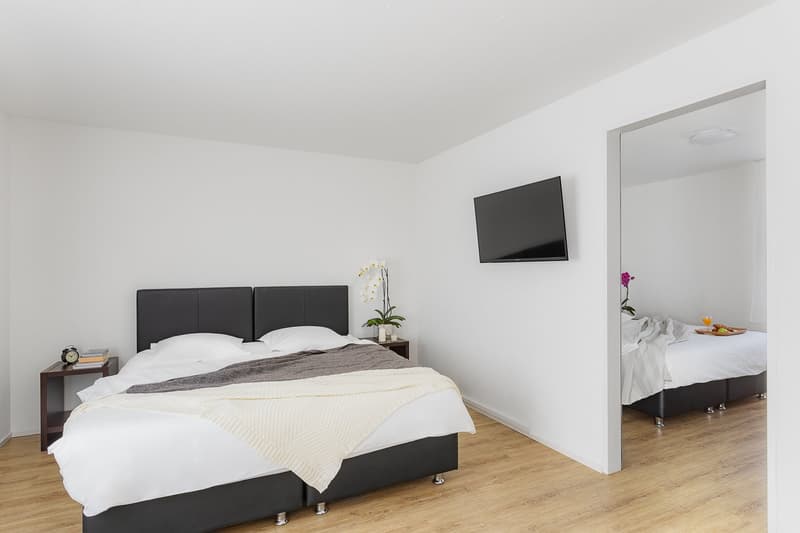 Two twin-sized beds, white bedding, blanket, TV on the wall, small nightstands, flowers, clean and tidy
