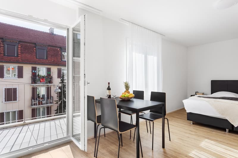 modern bedroom, glass door with a balcony, dining table with chairs, wine and fruit basket on the table, hardwood floors, white walls, clean white bedding on bed