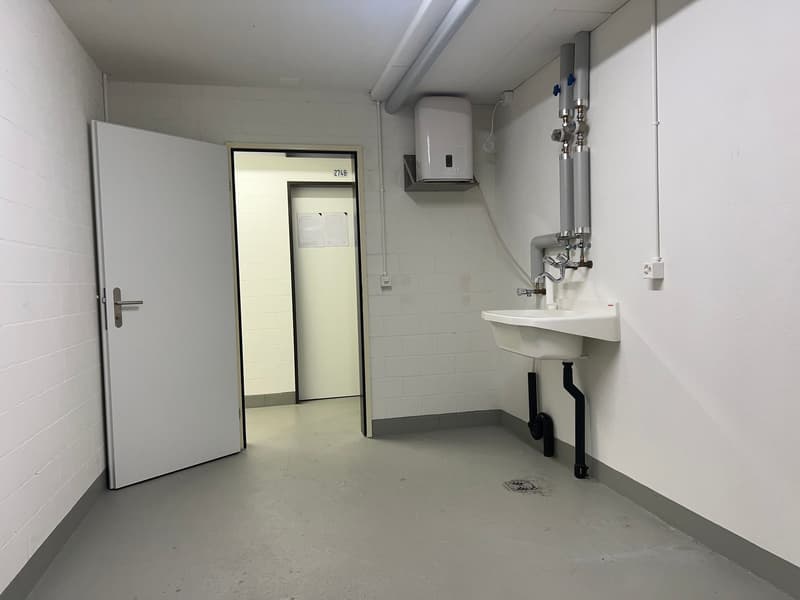A room with white walls, tiled floor, an open door, a sink with faucet, and pipes and fixtures mounted on the wall.