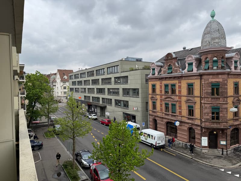 A street view of a city with buildings on both sides, parked cars, and people walking on the sidewalk.