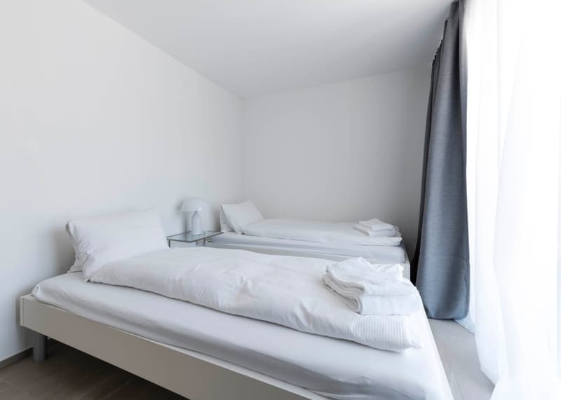 Spacious room with two beds, white walls, minimalist design, two lamps, bedside tables, folded towels on the beds.