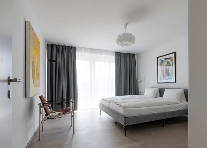 modern bedroom with bed, bed sheets, pillows, and bedside lamp, framed artwork, gray curtains, single wall painting, and modern chair