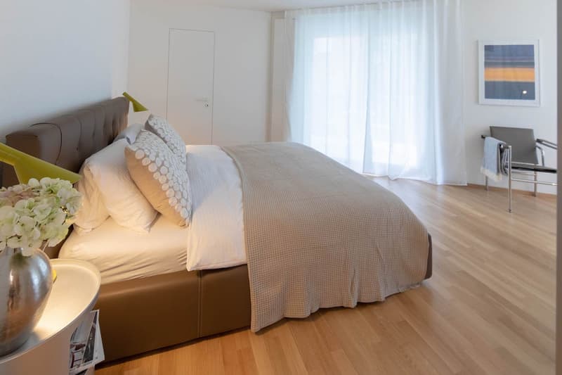 large bed, light wooden floors, white walls, white bedcover, pillows, two lamps, vase with flowers, chair, painting, curtain