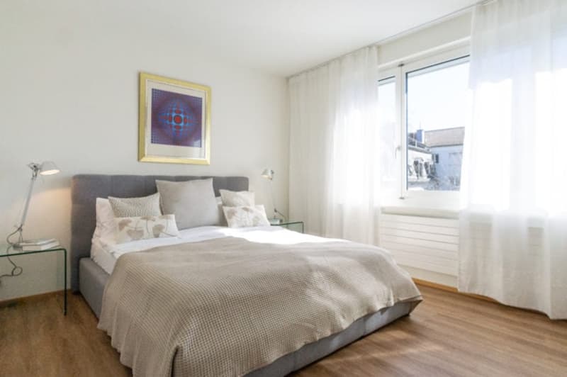 Double bed, gray headboard, white walls, wooden floor, two nightstands, white linens, two white lamps, white curtains, picture frame, radiator
