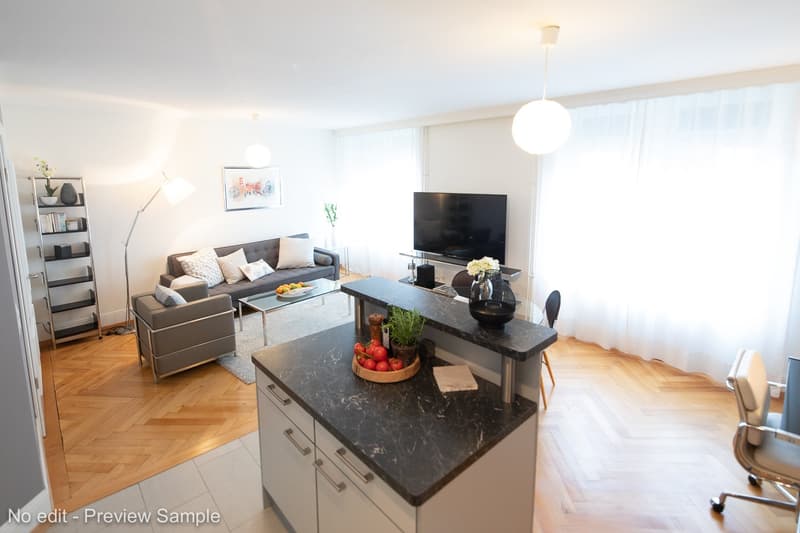 Spacious living room with hardwood floors, white walls, large windows, gray sofa, coffee table, TV stand, and kitchen island with black granite countertop and dishwasher