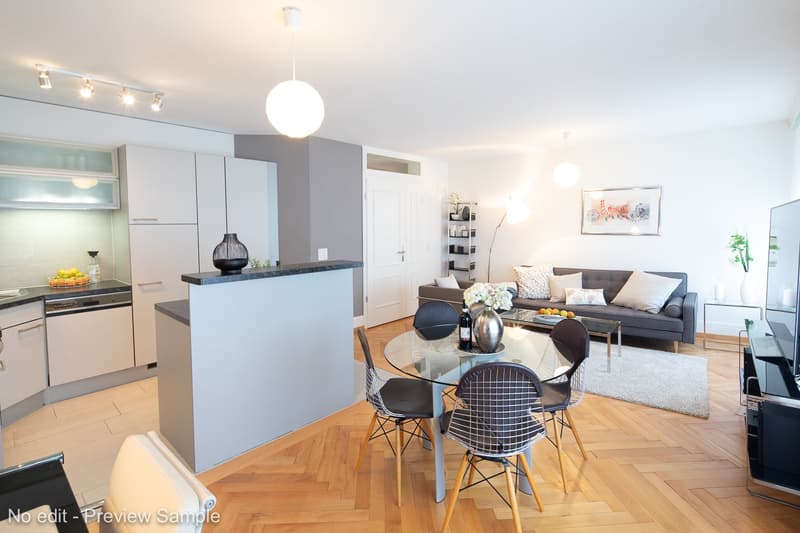 The image shows an open-plan living room and kitchen area. The kitchen features modern gray cabinets and a black countertop. There is a glass dining table with wire chairs in the center of the room. The living room has a gray sofa, a coffee table, and som