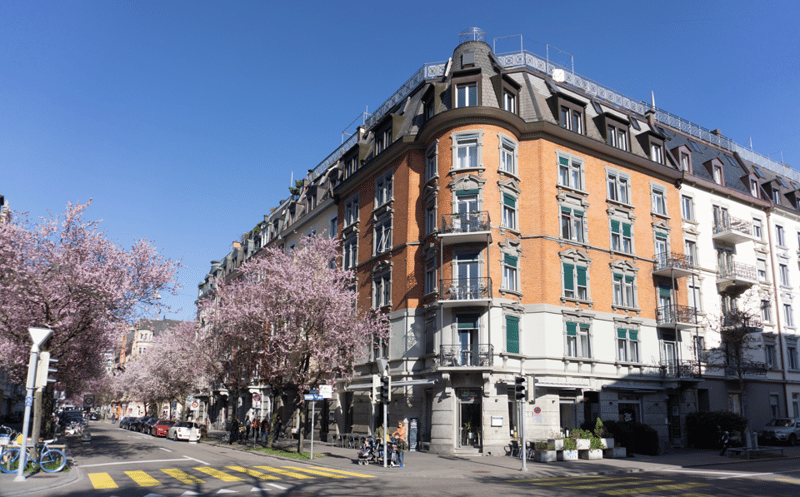 multistory building, orange facade, many windows, balconies, cherry blossoms in front, cars parked on street