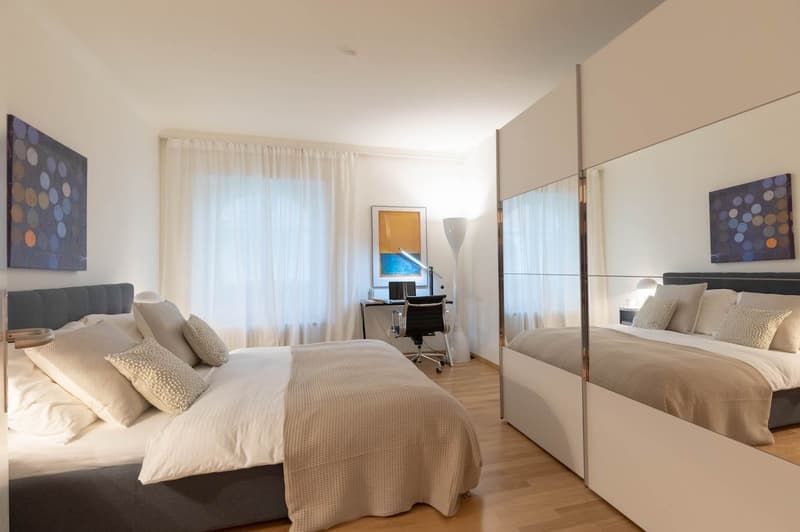 Bedroom with two beds, wooden floor, white walls, desk with monitor, and decorative artworks.