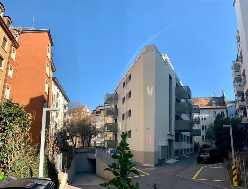 Modern apartment building with a raised ground floor, multiple balconies, and a driveway with parking spots